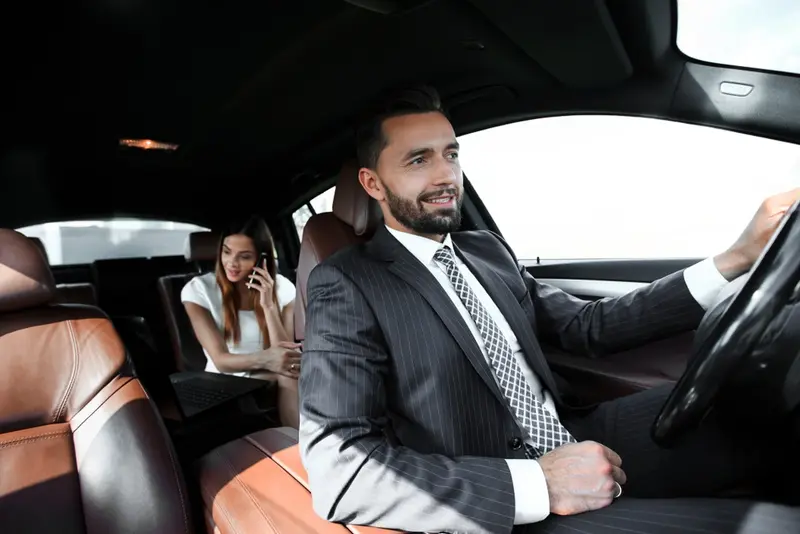 A chauffeur drives a luxurious black car, and in the back seat sits an elegant businesswoman talking on her phone, representing the luxury black car service in Austin.